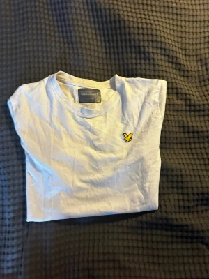 Vit t-shirt från Lyle & Scott - Vit t-shirt från Lyle & Scott med klassisk passform och korta ärmar. T-shirten har en rund halsringning och den ikoniska gula loggan broderad på bröstet. Perfekt basplagg i mjuk bomull.