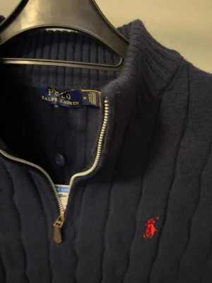 Mörkblå kabelstickad tröja Polo Ralph Lauren - Mörkblå kabelstickad tröja från Polo Ralph Lauren med halv dragkedja och hög krage. Tröjan har det klassiska röda Polo-logotypen broderad på bröstet och är tillverkad i ett mjukt, tjockt material. Perfekt för lager på lager under kyliga dagar.