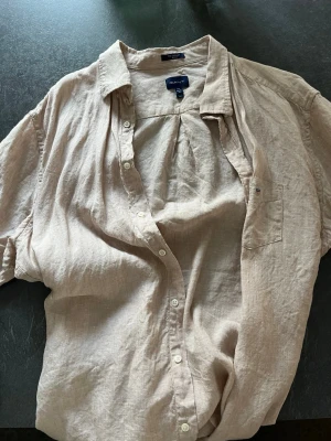 Beige linneskjorta från Gant - Snygg beige skjorta från Gant i luftigt linne. Skjortan har klassisk krage, kort ärm och knäppning framtill. Perfekt för en avslappnad och stilren look. Bröstficka med diskret logga på vänster sida.