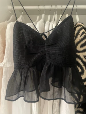 Svart volang linnetopp med tunna band - Supersöt svart linnetopp med tunna axelband och volangkant nedtill. Toppen har smockad överdel och transparent volang i nederkant som ger en luftig och trendig look. Perfekt för varma dagar eller som layering-piece. Storlek M men som en S/xs