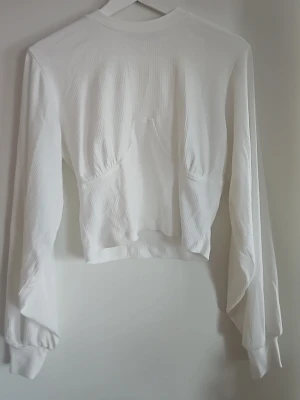 Vit ribbad långärmad topp - Vit långärmad topp med ribbad struktur och lätt croppad passform. Toppen har en unik sömdetalj framtill som ger en cool look. Perfekt att styla med jeans eller kjol.