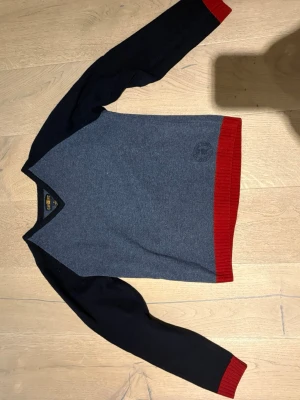 Blå stickad tröja från Gant - Snygg stickad tröja från Gant i blått med mörkblå ärmar och detaljer samt röda ribbade muddar vid ärmslut och nederkant. V-ringad hals och diskret logga på bröstet. Perfekt för dig som gillar klassisk men modern stil.