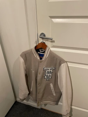 Beige och vit bomberjacka med brodyr - Snygg beige och vit bomberjacka i klassisk college-stil med broderad bokstavslogga på bröstet från Jack and Jones. Jackan har vita ärmar, ribbade muddar och två fickor framtill. Perfekt för dig som gillar streetwear och sportig look.