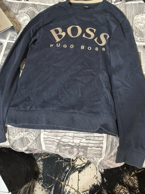 Mörkblå sweatshirt från Hugo Boss - Snygg mörkblå sweatshirt från Hugo Boss med stor BOSS-logga i beige framtill. Tröjan har rund halsringning, långa ärmar och ribbade muddar vid ärmslut och nederkant. Perfekt för en avslappnad och trendig stil.