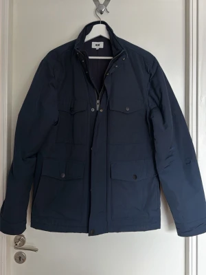 Mörkblå parkas från WE, storlek M - Stilren mörkblå parkas från WE med fyra praktiska fickor framtill och hög krage. Jackan har dragkedja och tryckknappar, perfekt för kyliga dagar. Materialet är slitstarkt och ger en clean look som funkar till det mesta.