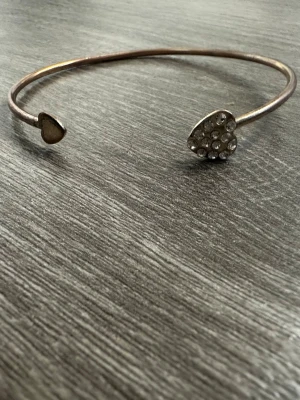 Guldfärgat armband med hjärtan - Snyggt öppet armband i guldfärgad metall med två hjärtformade detaljer, varav ena är dekorerad med glittrande stenar. Perfekt accessoar för att lyfta din outfit med lite extra bling och romantisk touch. Färgen har typ gått bort därav lågt pris, PRISET GÅR ATT DISKUTERA!!