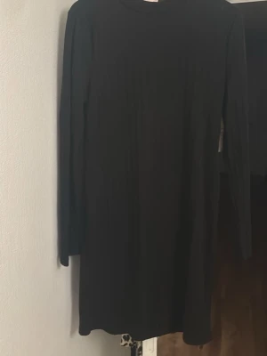 Svart ribbad klänning från Nelly - En svart ribbad klänning från Nelly med lång ärm och rund halsringning. Klänningen har en enkel och stilren design som passar perfekt till olika tillfällen. Materialet är mjukt och stretchigt för en bekväm passform.