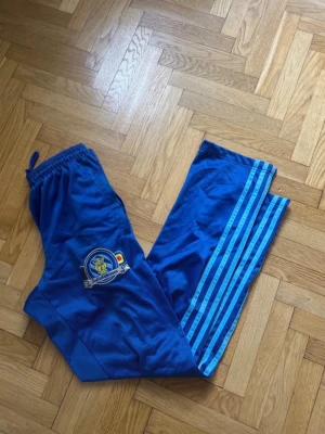 Blå träningsbyxor från sofialäktaren - Säljer ett par blå träningsbyxor med ljusblå ränder längs benen och ett broderat klubbemblem på ena låret. Byxorna har resår i midjan med dragsko och är tillverkade i ett lätt och bekvämt syntetmaterial, perfekta för träning eller uppvärmning.sjukt sällsynta byxor såldes för ett par år sedan.passar M