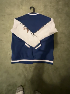 Blå collegejacka med vita detaljer - Snygg blå collegejacka med vita ärmar och tryck. Jackan har vita muddar och knappar framtill samt text och grafik på bröstet och ena ärmen. Perfekt för en sportig och avslappnad look.