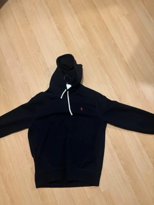 Svart hoodie från Polo Ralph Lauren - Svart hoodie från Polo Ralph Lauren med klassisk huva och vit dragsko. Liten röd broderad logga på bröstet och stor känguruficka framtill. Mjuk bomullsblandning som är skön att bära. Perfekt för en avslappnad och stilren look. Storlek L och hoodien är i väldigt fint skick.