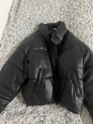 Svart cropped pufferjacka i skinnimitation - Säljer en svart pufferjacka i skinnimitation med hög krage och dragkedja framtill. Jackan har två snedställda fickor och quiltade detaljer som ger en cool och trendig look. Perfekt för dig som vill ha en edgy vinterstil.