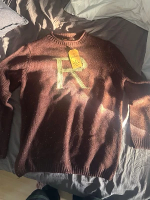 Brun Harry Potter stickad tröja - Aldrig använd, Stickad brun tröja från Harry Potter-kollektionen med ett stort 'R' på bröstet. Tröjan har rund hals och långa ärmar, perfekt för dig som älskar magi och vill ha en mysig och unik look.