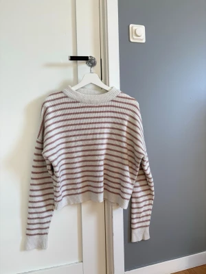 Randig stickad tröja från NA-KD - Mysig stickad tröja med ränder i vitt och beige. Tröjan har en avslappnad passform, ribbstickad rund halsringning samt muddar vid ärmslut och nederkant. 