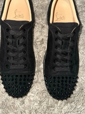 Svarta Louboutin sneakers med nitar - Säljer ett par svarta sneakers från Christian Louboutin med mattsvarta nitar på tån och klassisk röd sula. Skorna är låga och har ovandel i mocka med svarta skosnören. Perfekta för dig som vill sticka ut med en edgy och lyxig stil.