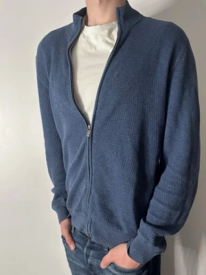 Blå ziptröja med ståkrage - Säljer en blå ziptröja med ståkrage och hel dragkedja. Tröjan har en diskret stickad struktur, långa ärmar och ribbade muddar vid ärmslut och nederkant. Perfekt för lager-på-lager och enkel att matcha med jeans eller t-shirt.