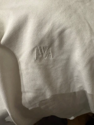 Vit pikétröja från JW Anderson - Klassisk vit pikétröja från JW Anderson med diskret broderad logga på bröstet. Tröjan har krage, knappar framtill och gröna detaljer på krage och ärmslut. Tillverkad i bomull för en skön och luftig känsla.