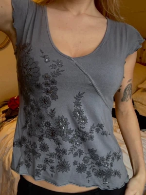 Grå blommig topp med paljettdetaljer - Grå topp med v-ringning och korta ärmar. Toppen har ett blommigt mönster framtill med svarta och silvriga paljettdetaljer. Materialet känns mjukt och toppen har en lätt vågig kant nertill. Väldigt gullig topp som funkar både för fest och vardag🌸
