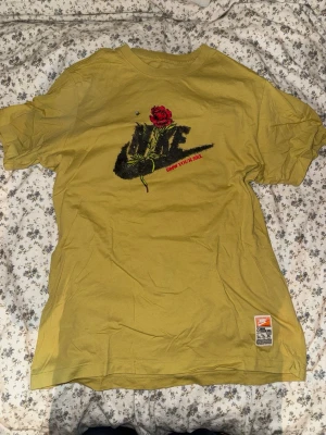 Gul Nike t-shirt med ros-tryck - Säljer en gul t-shirt från Nike med ett stort svart logotryck på bröstet och en röd ros ovanpå. Texten 'GROW YOUR SOLE' står i rött under loggan. Klassisk passform och rund hals. Liten etikett nertill. 