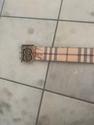 Burberry bälte med klassiskt rutmönster - Snyggt Burberry bälte med det ikoniska beigea och bruna rutmönstret samt en guldig TB-logga som spänne. Bältet har en exklusiv känsla och passar perfekt till att lyfta vilken outfit som helst. Helt nytt 