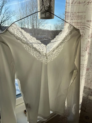 Vit långärmad topp med spets - Snygg vit långärmad loungewear-topp med djup V-ringning och dekorativ spetskant runt halsen. Tillverkad i mjukt och stretchigt material som känns skönt mot huden. Perfekt för chill dagar hemma eller som ett extra lager under tröjan.