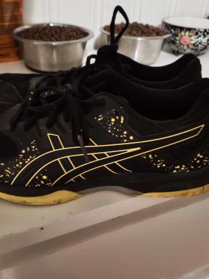 Svarta och gula Asics sneakers - Säljer ett par svarta Asics sneakers med gula detaljer och stänk på sidorna. Skorna har snörning och en låg profil med rund tå. Ovandelen är i mesh och syntetmaterial för bra andningsförmåga och komfort. Perfekta för dig som gillar sportig stil. Orginal sulan är bytt inne i skon 
