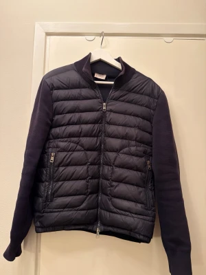 Svart pufferjacka från Moncler - Snygg svart pufferjacka från Moncler med quiltad front och tvåvägsdragkedja. Ärmarna är i stickad bomull vilket ger en cool kontrast till den glansiga, vadderade kroppen. Två fickor med dragkedja och ribbade muddar. Perfekt för dig som vill ha en stilren och modern jacka.