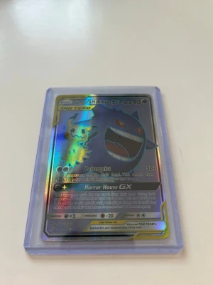 Pokémonkort Gengar & Mimikyu GX - Säljer ett holografiskt Pokémonkort med Gengar & Mimikyu GX TAG TEAM. Kortet har en skimrande yta med färger i regnbågseffekt och ligger i ett genomskinligt plastfodral. Perfekt för samlare av Pokémonkort eller TCG-fans.