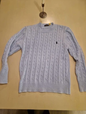 Ljusblå kabelstickad tröja Polo Ralph Lauren - Säljer en ljusblå kabelstickad tröja från Polo Ralph Lauren med klassisk rund halsringning och svart broderad logga på bröstet. Tröjan har ribbade muddar vid ärmslut och nederkant och är gjord i mjukt stickat material.