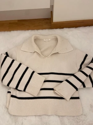 Vit stickad tröja med svarta ränder - Trendig stickad tröja i vitt med svarta horisontella ränder. Tröjan har en bred krage, långa ärmar och ribbade muddar samt nederkant. Perfekt till jeans för en avslappnad look.