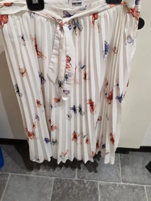 Vit plisserad kjol med fjärilsmönster Carolina gynning  - Vit långkjol från Carolina Gynning med plisserad design och färgglatt fjärilsmönster i rött, lila och blått. Kjolen har ett knytband i midjan för extra detalj och sitter snyggt med normal passform. Perfekt för dig som gillar en luftig och somrig stil.