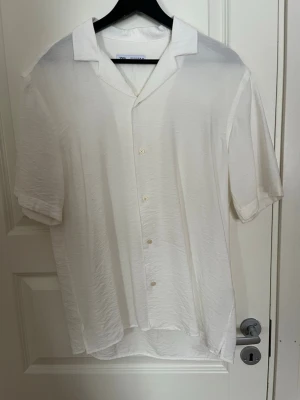 Vit kortärmad skjorta med knappar - Säljer en stilren vit skjorta med korta ärmar och klassisk krage. Skjortan har knappar framtill och är tillverkad i ett lätt, något strukturerat material som ger en avslappnad känsla. Perfekt för varma dagar och enkel att matcha med olika outfits.