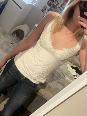 Vit spetsdetaljerad linnetopp - Snygg vit linnetopp med spetsdetaljer längs v-ringningen och axelbanden. Toppen har en figurnära passform och är ärmlös, perfekt för lager på lager eller sommarens varma dagar. Materialet känns mjukt och stretchigt.