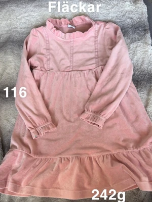 Rosa volangklänning med lång ärm - Söt rosa klänning med volangkant nedtill och långa ärmar. Klänningen har en hög krage med rynk och dekorativa sömmar framtill. Tillverkad i mjukt tyg, perfekt för en trendig och bekväm look.🌸 Fläckar. Storlek 116 