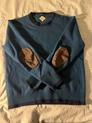 Blå stickad tröja från Boggi Milano - Mysig blå stickad tröja från Boggi Milano med mörkblå ribbade muddar och nederkant. Tröjan har bruna armbågslappar i kontrastmaterial och rund halsringning. Tillverkad i mjuk ull och designad i Italien. Perfekt för kalla dagar.