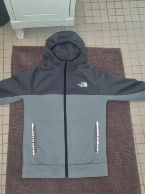 Grå hoodie från The North Face - Snygg tvåtonad grå hoodie från The North Face med hel dragkedja framtill och huva. Loggan är tryckt på bröstet och fickor med dragkedja finns på båda sidor. Perfekt för chill eller när du är på språng.