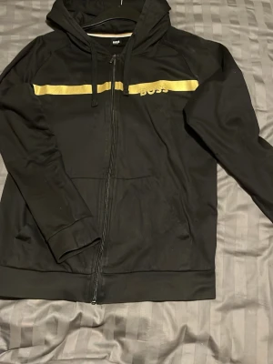 Svart hoodie med guldtryck från BOSS - Snygg svart hoodie från BOSS med dragkedja och huva. Framtill finns en guldfärgad rand och BOSS-logga i guld. Hoodien har dragsko i huvan och är tillverkad i mjukt material, perfekt för en chill och stilren look.