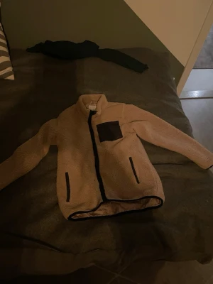 Beige teddyjacka med svarta detaljer - Mysig beige teddyjacka med svarta dragkedjor och en svart bröstficka. Jackan har hög krage och två sidofickor med dragkedja. Perfekt för kalla dagar och ger en trendig look.