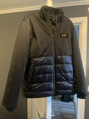 Svart pufferjacka från EA7 - Snygg svart pufferjacka från EA7 med glansig vadderad nederdel och slät ovandel. Jackan har hög krage, dragkedja framtill och EA7-logga på bröstet. Perfekt för kalla dagar och riktigt trendig stil.