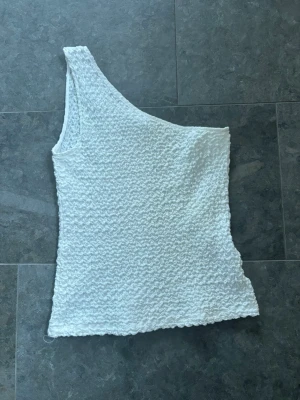 Vit oneshoulder topp i strukturmönster - Säljer en vit oneshoulder topp med snygg struktur i tyget. Toppen är ärmlös och har en unik asymmetrisk design som ger en cool vibe. Perfekt för dig som vill sticka ut lite extra. Materialet känns mjukt och stretchigt.