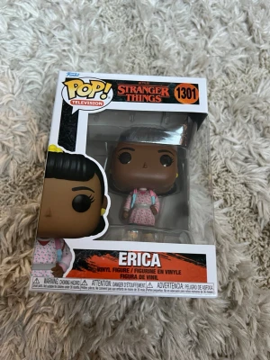 Funko Pop Stranger Things Erica - Säljer en Funko Pop vinylfigur av Erica från Stranger Things, nummer 1301. Figuren är färgglad med rosa kläder och står i originalförpackning med fönster. Perfekt för samlare eller fans av serien.