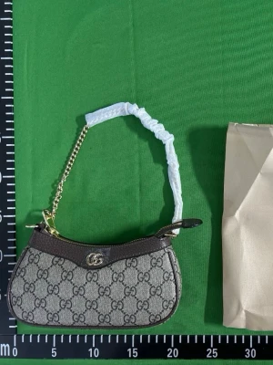 Gucci axelremsväska med GG-mönster - Säljer två snygga axelremsväskor från Gucci med klassiskt GG-monogram. Ena väskan är beige med bruna detaljer och guldfärgad kedja, den andra är ljusbeige med matchande detaljer. Båda har guldfärgad logga framtill och kort axelrem. Perfekt för dig som gillar lyxig och trendig stil. Finns även olika färger och modeller. 