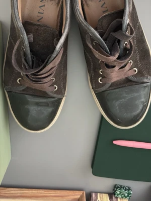 Bruna sneakers från Lanvin - Snygga bruna sneakers från Lanvin med mocka och canvas på ovandelen samt tåparti i svart gummi. Skorna har bruna skosnören och vita sulor. Perfekta för dig som gillar en stilren och klassisk look.