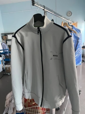 Grå ziptröja från BOSS - Snygg grå ziptröja från BOSS med svarta detaljer längs dragkedjan och axlarna. Tröjan har hög krage, två fickor framtill och en stilren logga på bröstet. Perfekt för en sportig och avslappnad look.