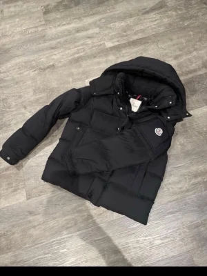 Svart dunjacka från Moncler - Snygg svart dunjacka från Moncler med huva och klassisk logotyp på ärmen. Jackan har dragkedja och knappar framtill samt två stora fickor. Perfekt för kalla vinterdagar och riktigt stilren look.