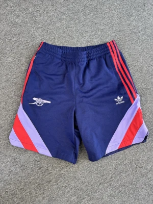 Adidas x Arsenal blå shorts - Snygga blå shorts från Adidas i samarbete med Arsenal, med röda och lila detaljer på sidorna samt klassiska Adidas-ränder. Elastisk midja och sportig look, perfekt för fotboll eller träning.