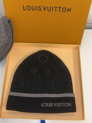 Svart monogram mössa  - Snygg svart mössa med klassiskt monogrammönster och grå kant. Mössan har en diskret logga broderad framtill och är tillverkad i mjukt, stickat material som håller dig varm och stilren.