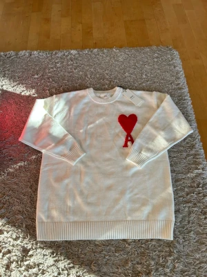 Vit stickad tröja från Ami Paris - Vit stickad tröja från Ami Paris med rund halsringning och ribbade muddar. På bröstet finns ett rött hjärta och bokstaven 'A' broderat. Tröjan har en avslappnad passform och är perfekt för en trendig vardagslook.