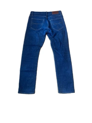 Mörkblå jeans från Tiger of Sweden - Snygga mörkblå jeans från Tiger of Sweden med klassisk femficksdesign och raka ben. Jeansen har kontrastsömmar och en brun läderpatch med logga bak i midjan. Perfekta för en stilren och avslappnad look.