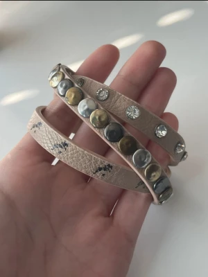 armband med nitar och stenar - Snyggt armband i läderimitation som lindas flera varv runt handleden. Dekorerat med runda nitar i silver och guld samt gnistrande klara stenar. Perfekt accessoar för att ge din outfit en cool touch. 