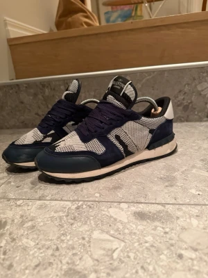 Valentino blåvita sneakers - Säljer mina riktigt snygga Valentino rockrunners  i blått och vitt med mesh- och mockadetaljer. Skorna har en låg siluett, vita och mörkblå partier samt mörkblåa snören. Perfekta för en trendig streetstyle-look. Mycket bra skick utan några defekter. Dustbag tillkommer. Strorlek 42 passar även 43. Skriv för fler bilder så löser jag. Pris kan diskuteras